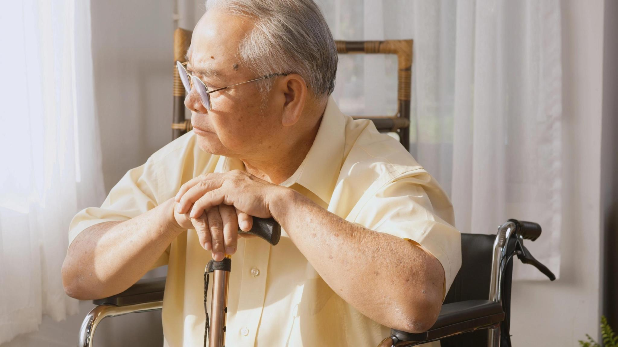 An elderly man in a wheelchair resting his hands on a cane, looking out a window