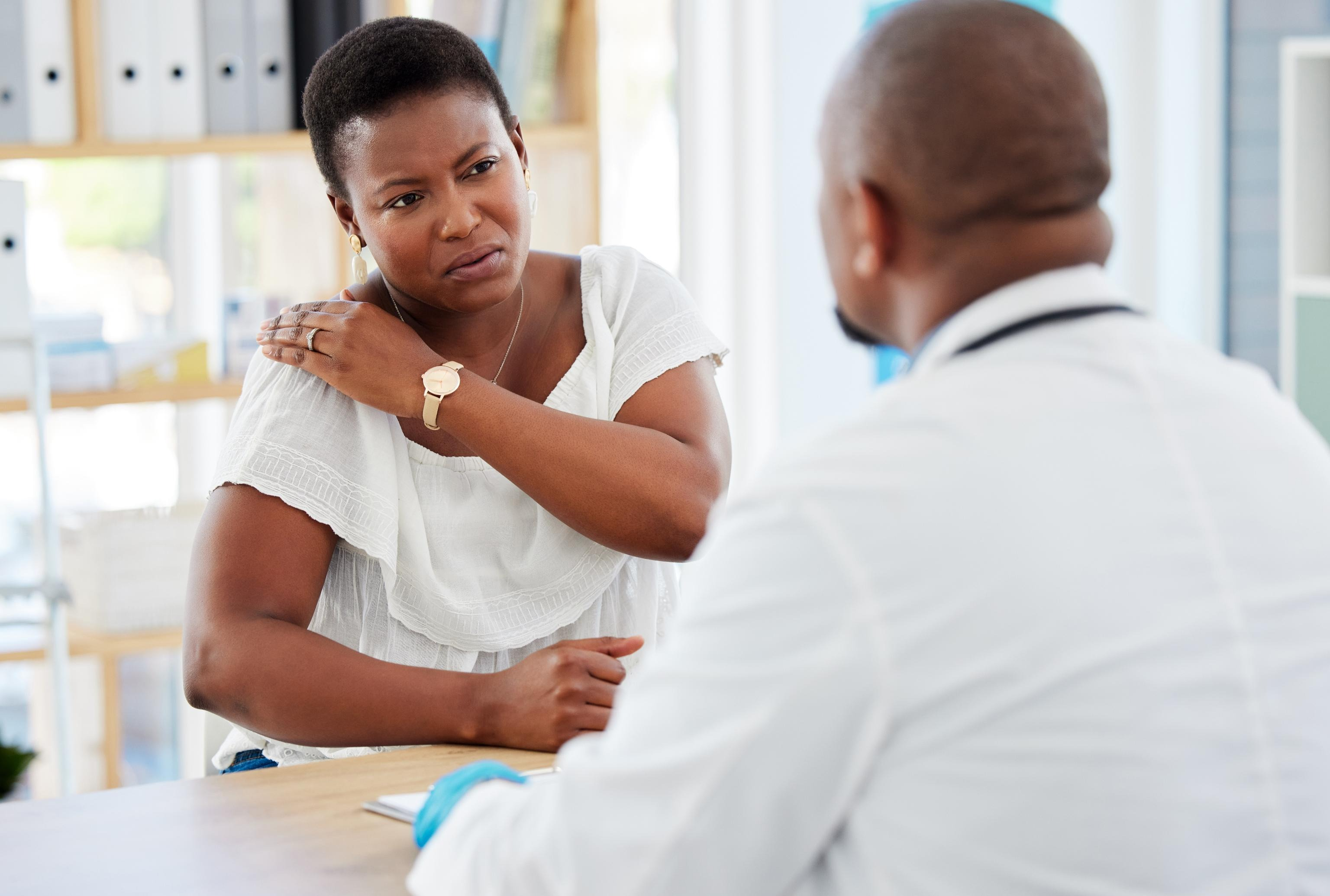 A woman holding her shoulder in pain while consulting with a doctor at his desk